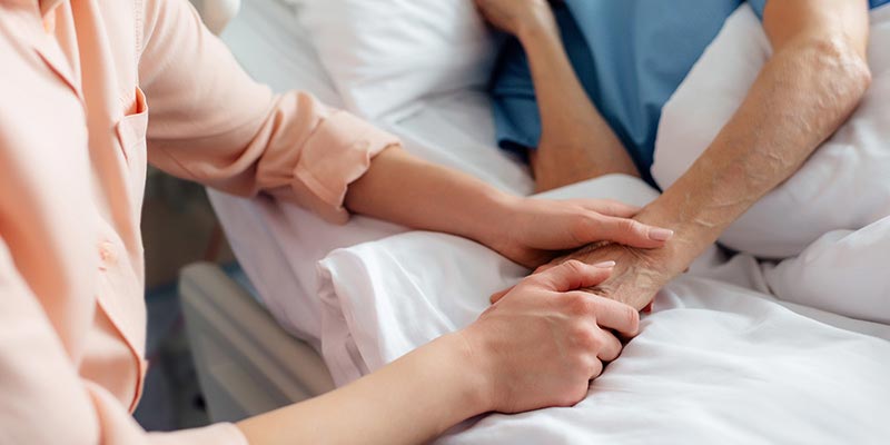 Woman holding the hand of an elderly lady in a hospital bed.