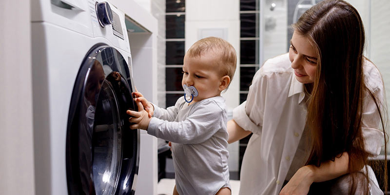 Young mum watching baby hold onto washing machine for support to stand.