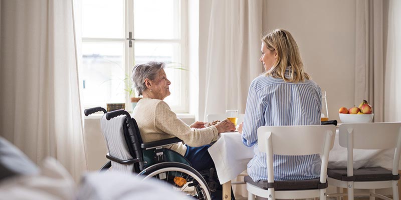 Young woman sitting at a table with an elderly woman, who is in a wheelchair.